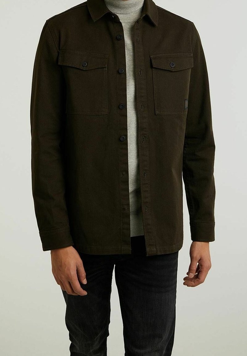 Homme portant une veste à boutons marron foncé sur un pull à col roulé gris clair et un pantalon noir, debout devant un fond uni.