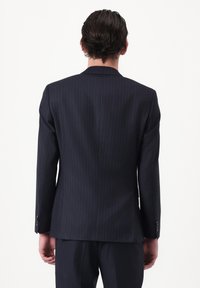 Homme portant un costume sombre à rayures fines avec une veste et un pantalon assortis, vu de dos sur un fond blanc uni.