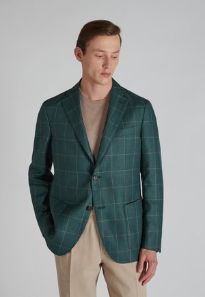 Türkiser Blazer mit einem Karomuster, zwei Fronttaschen und zwei Knöpfen. Getragen über einem beigen Pullover, kombiniert mit hellen Hosen.