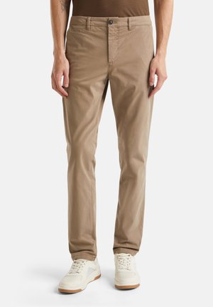 Beige Baumwoll-Chinos im Slim Fit mit Knopfverschluss und Fronttaschen, kombiniert mit weißen Sneakers vor einem neutralen Hintergrund.