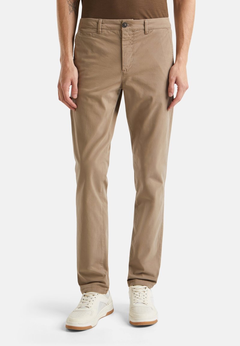 Beige Baumwoll-Chinos im Slim Fit mit Knopfverschluss und Fronttaschen, kombiniert mit weißen Sneakers vor einem neutralen Hintergrund.