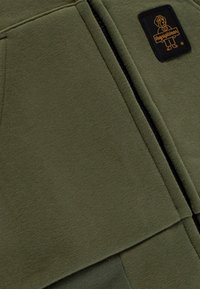 Pullover in pile verde oliva con chiusura a zip, accenti a coste e logo a patch nero con dettagli gialli. Texture morbida e design caldo.