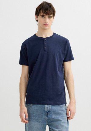 Joven hombre que lleva una camiseta henley de manga corta azul marino con tres botones y vaqueros de mezclilla azul claro, de pie frente a un fondo gris liso.
