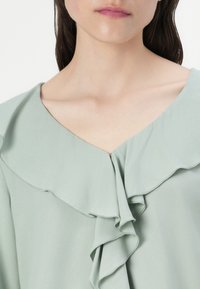 Lys grøn bluse med dyb V-hals, lagdelte flæsedetaljer, glat tekstur og en løs pasform omkring halsen.