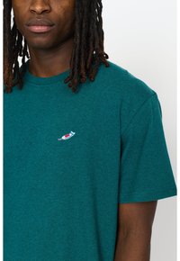 Teal katoenen T-shirt met ronde hals, voorzien van een kleine geborduurde afbeelding van een tandpastatube op de linkerkant van de borst.