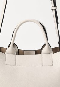 Sac tote en cuir blanc avec deux poignées arrondies, présentant une surface texturée et des bords contrastants noirs. L'intérieur est beige.