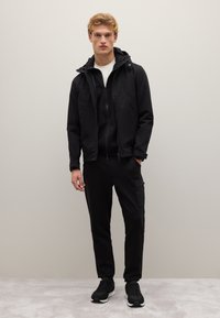 Schwarze Zip-up-Jacke mit Kapuze, kombiniert mit einem schwarzen Zip-up-Sweatshirt und Cargo-Hosen. Sneakers mit weißen Sohlen. Glatte, strukturierte Materialien.