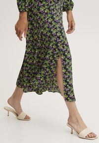 Vestito floreale con motivo verde, viola e nero, caratterizzato da uno spacco laterale. Abbinato a mule con tacco nude, che mostrano una texture liscia e una forma elegante.