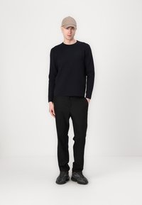 Marineblauer Pullover, schwarze Hose und schwarze Schuhe; das Model trägt eine beige Mütze. Einfaches Design, taillierte Form, glatte Textur.