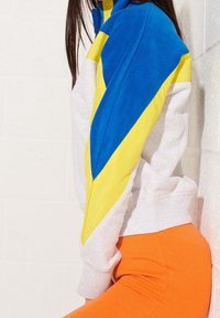 Osoba ubrana w kurtkę w kolorowe bloki, w niebieskie, żółte i białe sekcje, w parze z jasnopomarańczowymi spodniami, stojąca na tle białej ściany.