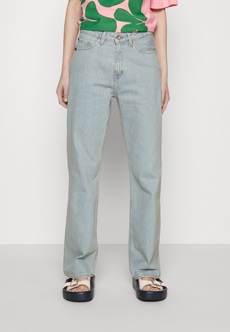 Jeans de pierna recta en azul claro con diseño de cinco bolsillos, que presentan un ligero desgaste y una textura clásica de denim; combinados con una blusa de patrón rosa y verde.