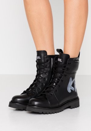 Bottes de combat noires à lacets avec semelles épaisses et lettrage gris style graffiti sur le côté extérieur portées sur des jambes nues.