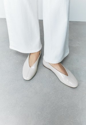 Person steht auf grauem Boden, trägt weiße Hosen mit weitem Bein und glänzende beige flache Schuhe mit V-förmiger Öffnung.