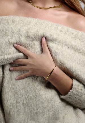 Pulsera dorada sobre una mano descansando en un suéter de punto texturizado color crema. La tela parece suave y gruesa.