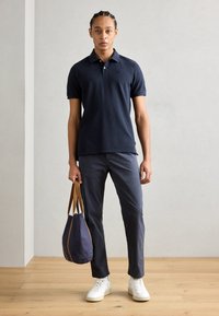 Marinblå polo med korta ärmar, kombinerad med mörkgrå byxor och vita sneakers. Håller en marinblå väska med bruna handtag.