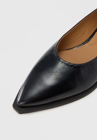Chaussure noire en cuir à bout pointu avec une texture lisse et un design ajouré. Elle possède une semelle plate noire et une semelle intérieure beige.