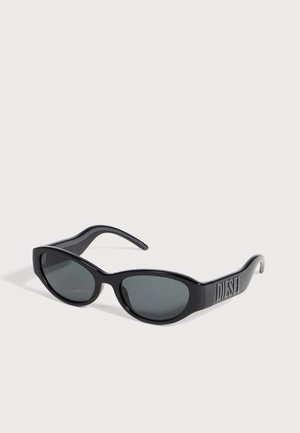 Lunettes de soleil Diesel noires, avec des verres ovales gris et des lettres en relief sur les branches larges, sur fond clair.
