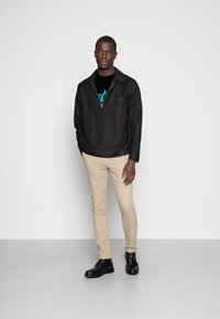 Schwarze Zip-Jacke mit Kragen, Tasche, schwarzes T-Shirt mit blauem Grafikdesign, beiger Slim-Fit-Hose, schwarze Schuhe, stehend vor einem hellen Hintergrund.