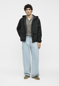Jeune homme portant une veste noire à capuche, un polo gris, un jean large bleu clair et des baskets vertes, debout devant un fond blanc.