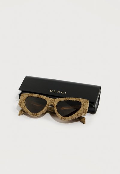 Lunettes de soleil Gucci à motif marron avec verres foncés reposant devant un étui à lunettes noir de marque Gucci sur une surface blanche.