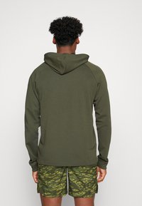Olivgrüner Kapuzenpullover mit strukturierter Oberfläche und Raglanärmeln, kombiniert mit gemusterten gelb-grünen Shorts.