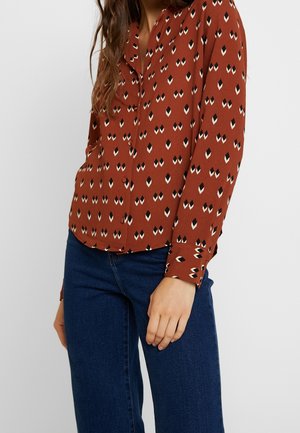 Femme portant une blouse boutonnée couleur rouille avec un motif en losanges noir et blanc, associée à un jean bleu foncé sur un fond uni.