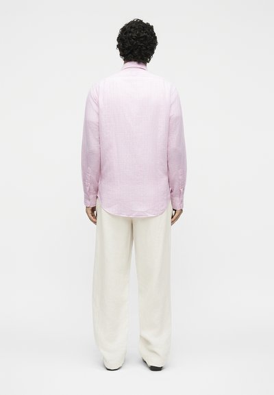 Ενήλικας με κατσαρά μαλλιά στέκεται με το σώμα γυρισμένο, φοράει ένα ροζ μακρυμάνικο πουκάμισο και άνετο λευκό παντελόνι σε απλό φόντο.