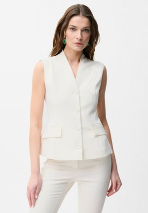 Woman wearing sleeveless white blazer with buttons and matching white trousers, accessorized with green drop earrings.