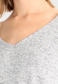 Grauer strukturierter Pullover mit V-Ausschnitt, der ein weiches, leicht marmoriertes Muster aufweist. Der Stoff scheint leicht und atmungsaktiv zu sein.