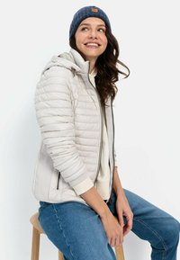 Lynlét skumjakke i crème med lynlås fortil, ribbede manchetter og hætte; båret over en crème sweater og parret med blå jeans og en marineblå hue.