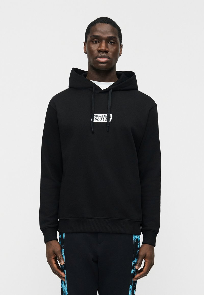 Jeune homme debout, portant un sweat à capuche noir avec le texte "Versace Jeans Couture" et un pantalon noir avec des rayures latérales à motifs bleus.