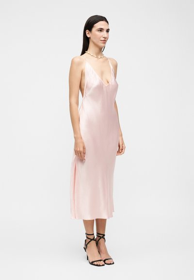 Γυναίκα με μακριά σκούρα μαλλιά που φοράει ανοιχτό ροζ σατέν φόρεμα με λεπτές τιράντες και μαύρα ψηλοτάκουνα σανδάλια με λουράκια, στέκεται μπροστά σε λευκό φόντο.