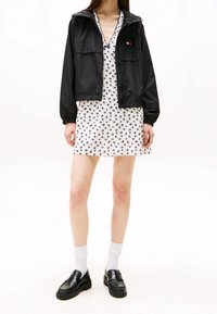 Czarna kurtka zapinana na zamek błyskawiczny z dwoma kieszeniami z przodu, na białej sukience z niebieskim wzorem ptaków. Modelka ma na sobie czarne mokasyny i białe skarpetki.