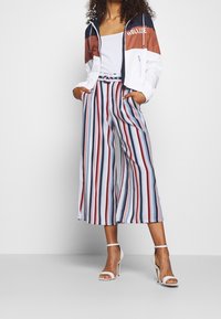 White cropped jacket with color-blocked sections in navy and rust, over white tank top. Striped wide-leg pants in red, blue, and white. White heels.