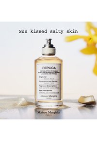 Butelka perfum oznaczona Replica by Maison Margiela na piasku z muszlą i żółtym kwiatem, tekst brzmi: Skóra muśnięta słonym słońcem.