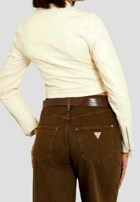 Woman wearing a cropped cream jacket and high-waisted brown pants with a brown textured belt, shown from the back against a plain background.