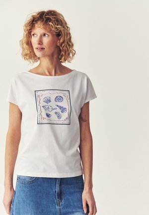 Kobieta o kręconych włosach nosi białą koszulkę z niebieskimi i czerwonymi ilustracjami muszli, połączoną z niebieską spódnicą dżinsową, na jednolitym tle.