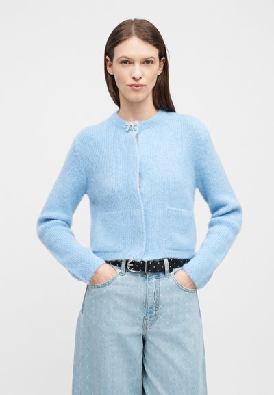 Γυναίκα με μακριά καστανά μαλλιά φοράει ένα ανοιχτό μπλε μάλλινο ζακέτα, μπουκαρισμένη σε ψηλόμεσα ανοιχτό μπλε τζιν με μαύρη ζώνη, τα χέρια στις τσέπες.