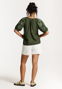 Blouse vert olive avec des manches bouffantes et des détails floraux brodés, associée à un short blanc et des sandales vert clair. Texture à pois présente.