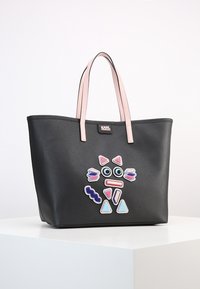 Svart tote-väska med ljusrosa handtag som har färgglada abstrakta ansiktsformade lappar på framsidan och en liten varumärkeslogotyp.