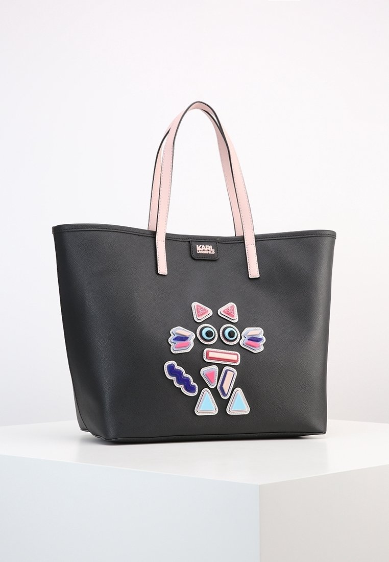 Svart tote-väska med ljusrosa handtag som har färgglada abstrakta ansiktsformade lappar på framsidan och en liten varumärkeslogotyp.