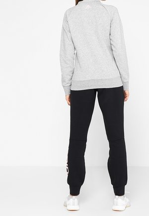 Woman standing, wearing a light gray sweatshirt, black sweatpants with a side logo, and white sneakers, shown from the back.