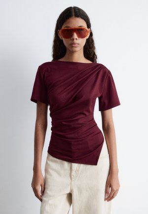 Burgundy short-sleeve top with a draped design, featuring a boat neckline and asymmetrical hem, paired with orange sunglasses.