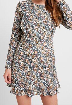 Femme portant une robe à manches longues à imprimé floral avec des volants sur l'ourlet et les épaules, debout devant un fond uni.