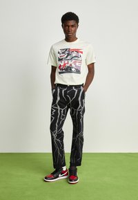 T-shirt graphique jaune clair avec un design rétro, pantalon noir avec des motifs d'éclairs blancs, et baskets noires/rouges.