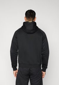 Μαύρο φούτερ με κουκούλα από υφή υφάσματος, με κορδόνι στην κουκούλα και λογότυπο VENUM σε κίτρινο στο πίσω μέρος. Συνδυασμένο με μαύρο σορτς με σχέδια.