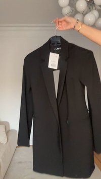 Schwarzer Blazer mit langen Ärmeln, Ein-Knopf-Verschluss und Revers mit Kerbe. Der Stoff wirkt glatt und leicht strukturiert. Mit Etikett versehen.
