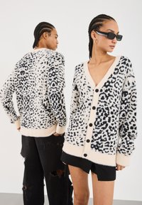 Fluffy cream cardigan with black leopard spots, featuring a V-neck, long sleeves, and black buttons down the front. Textured, cozy design.