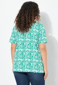 Zelené tričko s krátkým rukávem s bílým květinovým vzorem, vyrobené z měkké tkaniny s volným střihem. Pozadí je čistě bílé.