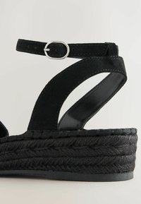 Zwarte wedge-sandaal met een bovenwerk van suede, geweven textuur op het platform, enkelbandje met zilveren hardware en een lichte kromming in het design.